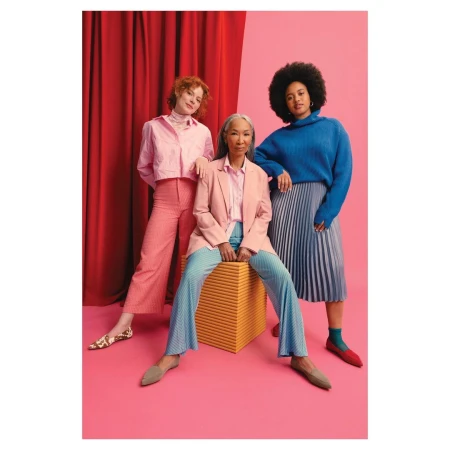 Three women of diverse ages pose in a colorful studio setting, wearing pink and blue attire with a pink and red backdrop.