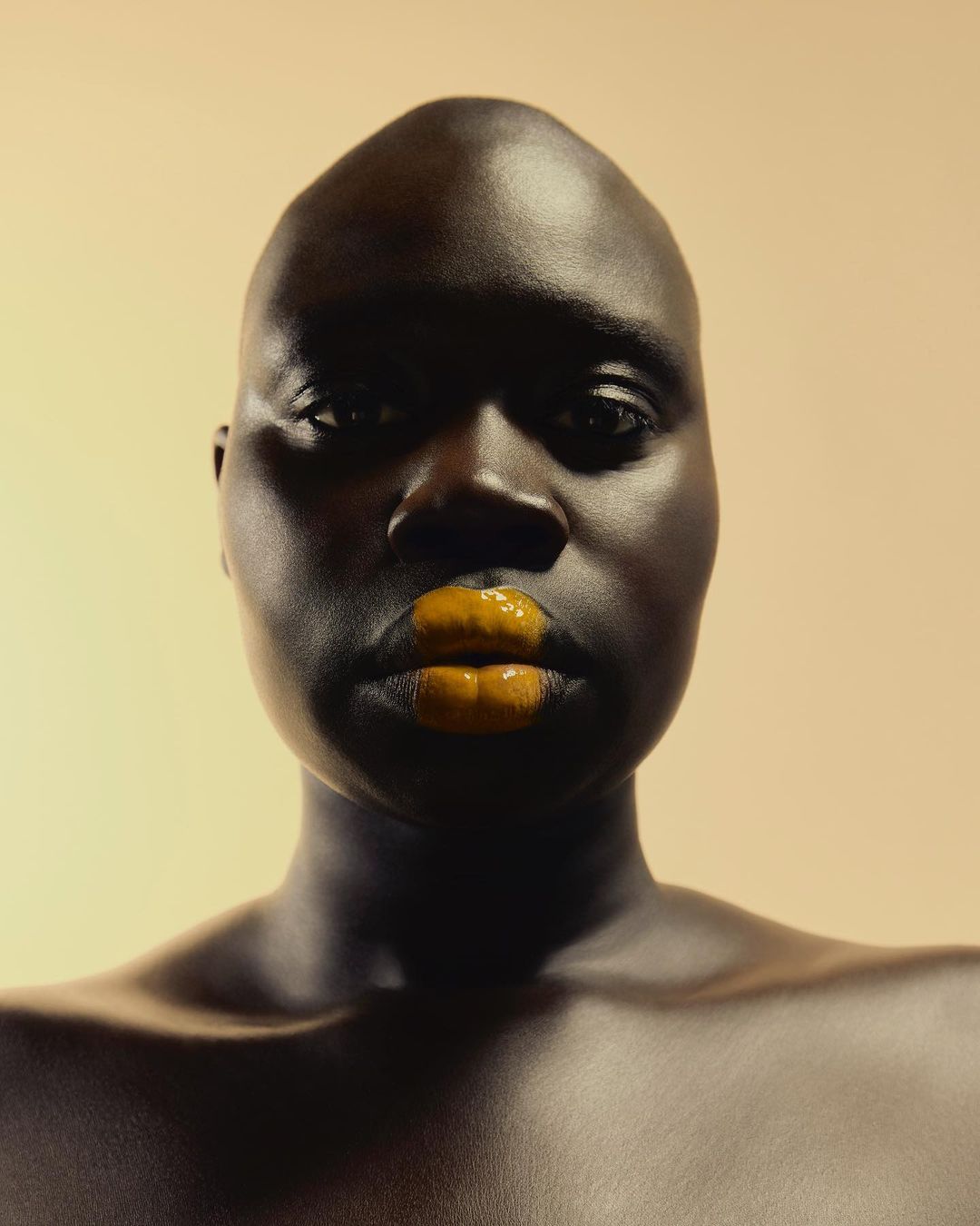 Close-up studio portrait of a bald adult with deep skin tone and glossy amber lips, looking directly into the camera on a neutral backdrop.