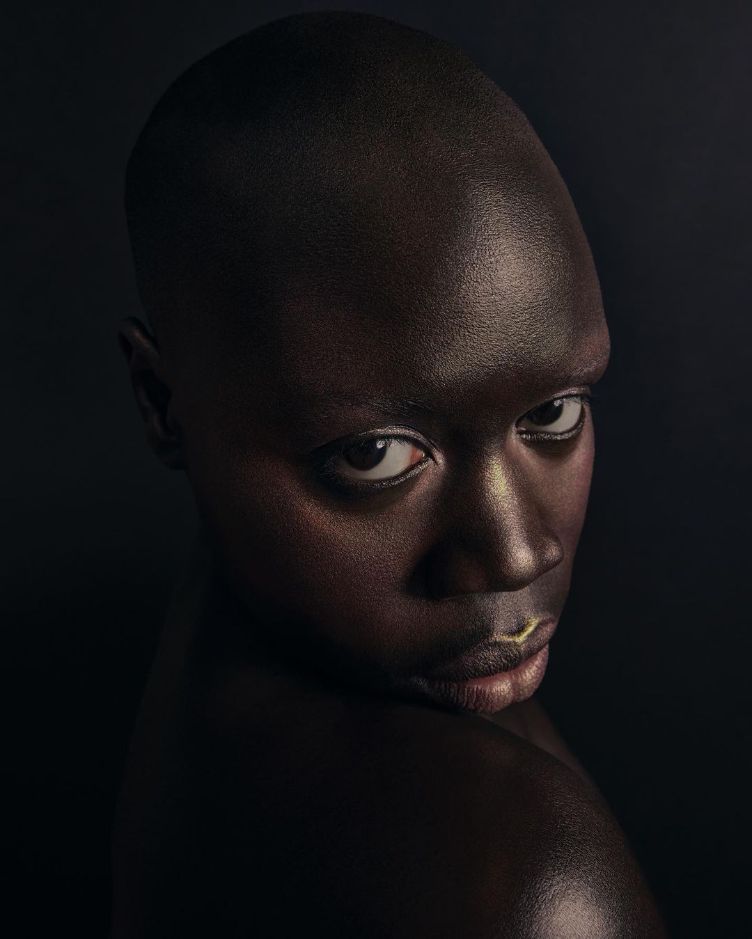 Portrait of an African woman with a shaved head looking over her shoulder in a dark studio with cool lighting and gold lip highlights.