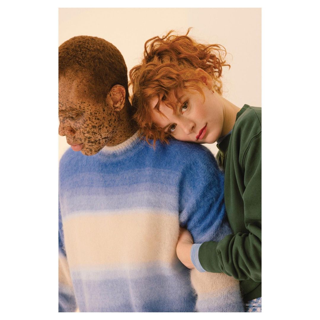 Two fashion models pose indoors against a cream-colored background. One model rests her head on the shoulder of the other model.
