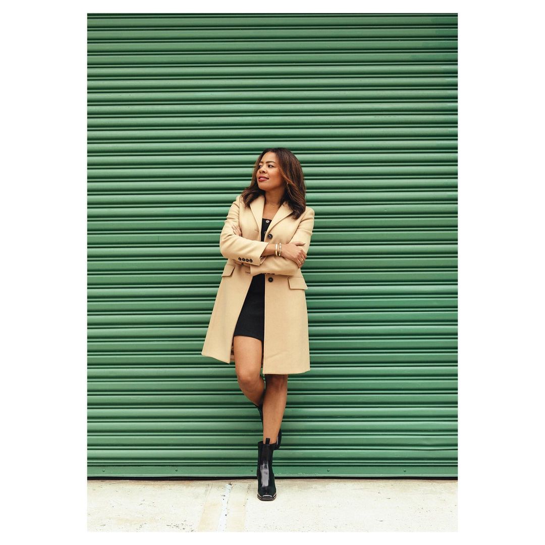 A stylish woman in a tan coat stands confidently against a green corrugated metal wall wearing a black dress and boots, looking aside.