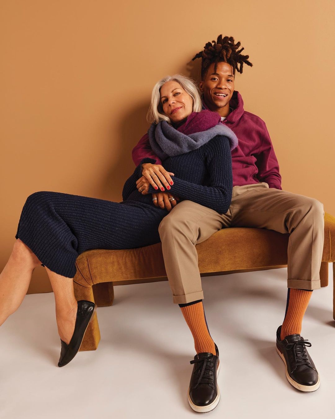 Mature woman in dark dress and Black man in maroon hoodie sit on bench in tan studio, smiling, hugging, in a fashion portrait.