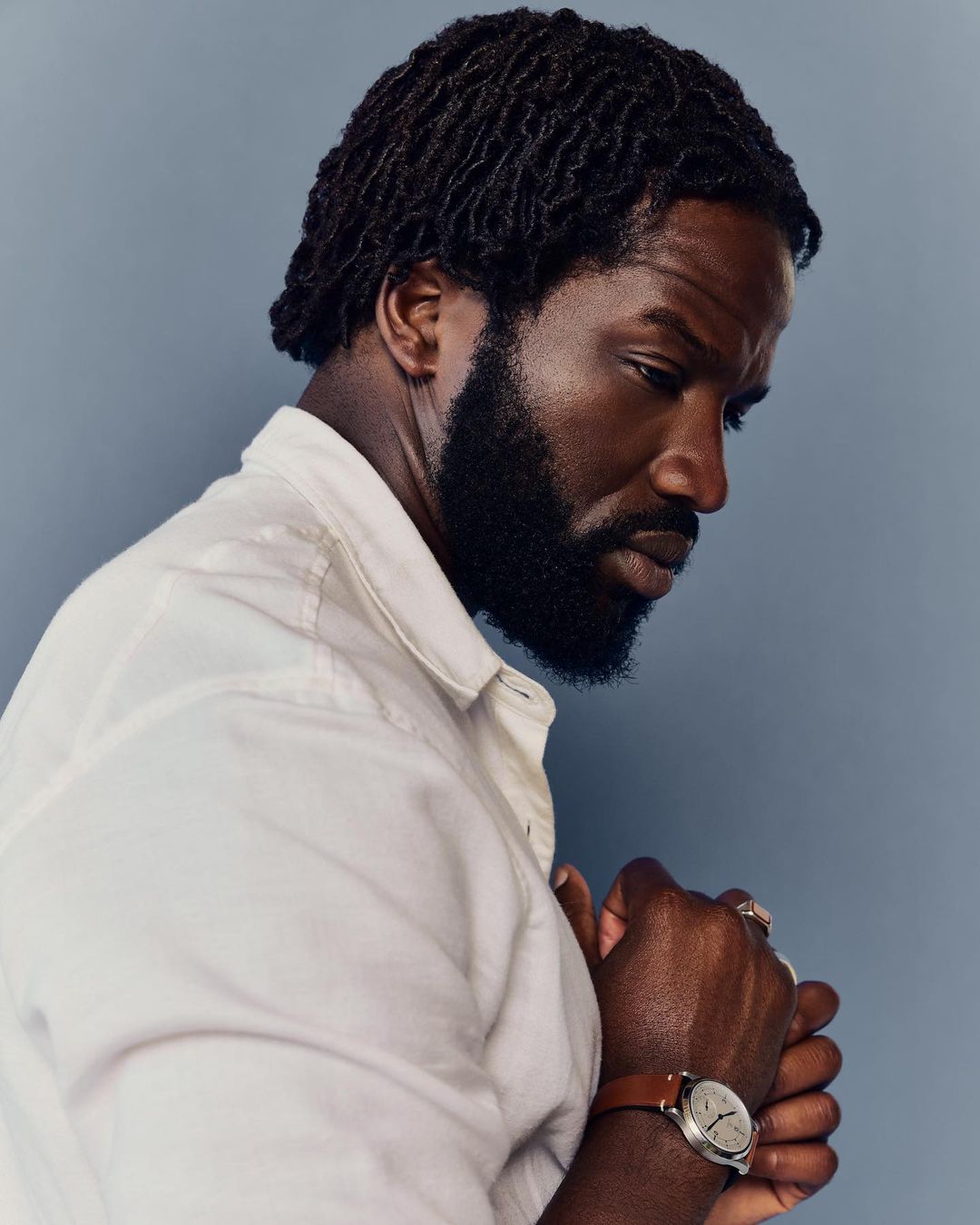 Profile portrait of a black adult male with dreadlocks and a beard wearing a white shirt and a watch against a blue background.