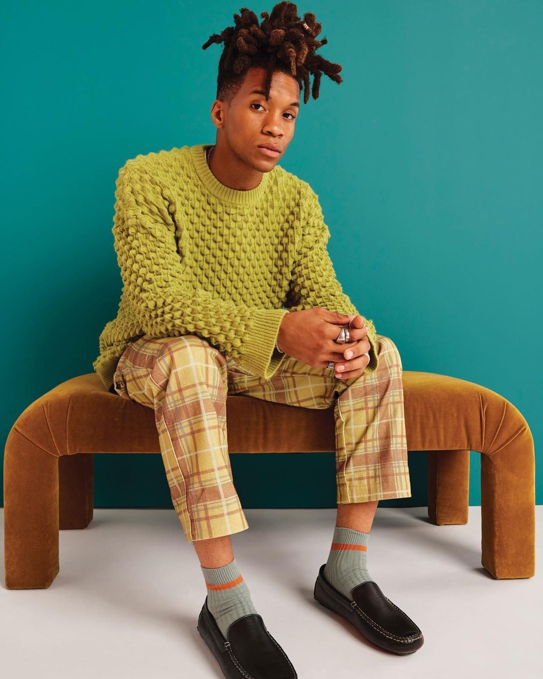 Young adult male posing on a brown bench against a blue wall, wearing a green sweater, plaid pants, socks, and loafers indoors.
