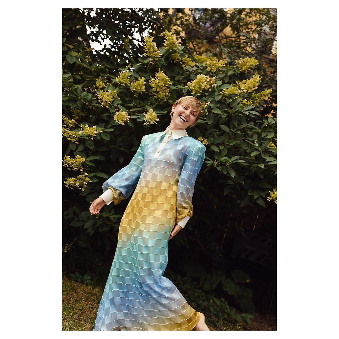 Smiling woman in a long blue and yellow checkered dress posing in front of lush green foliage and yellow flowers