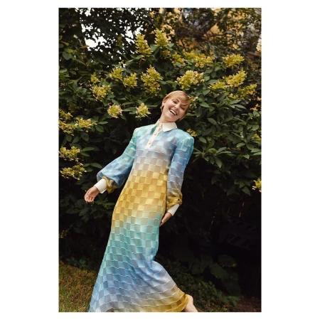 Smiling woman in a long blue and yellow checkered dress posing in front of lush green foliage and yellow flowers