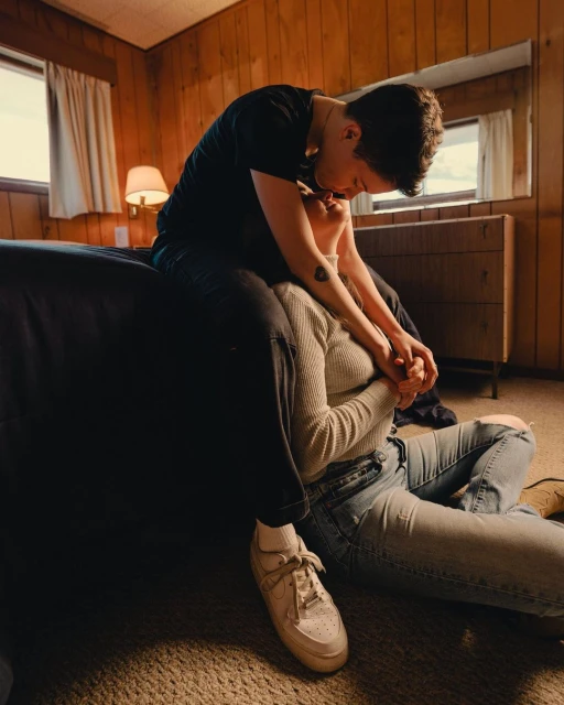 Two adult women in an embrace inside a wood-paneled bedroom with neutral tones, one sitting on the other who is on the floor.