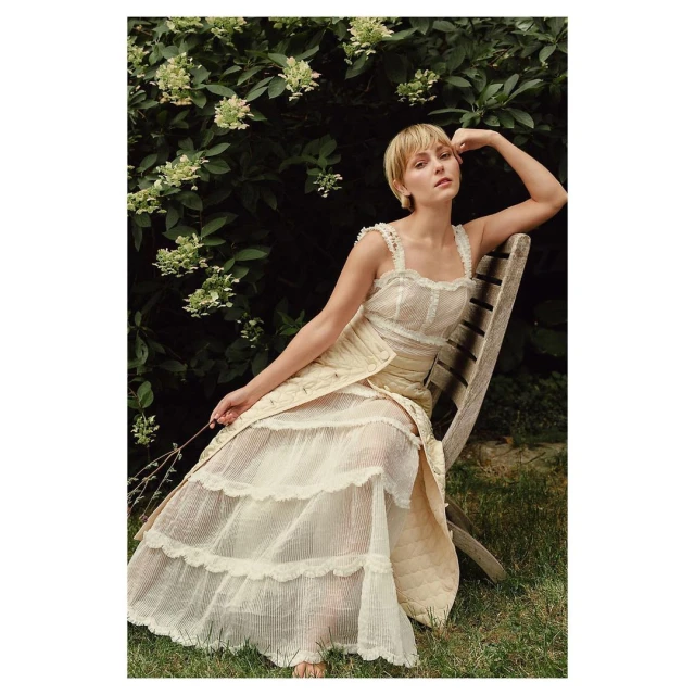 Adult female model with short blonde hair in a light-yellow semi-sheer dress posing on a wood chair outdoors against a green foliage background.