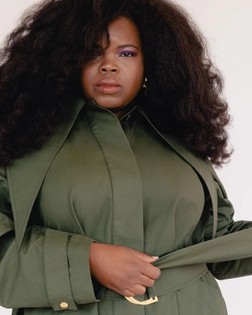 Headshot of an African-American woman with large hair and purple eyeshadow wearing an olive green trench coat with gold hardware.