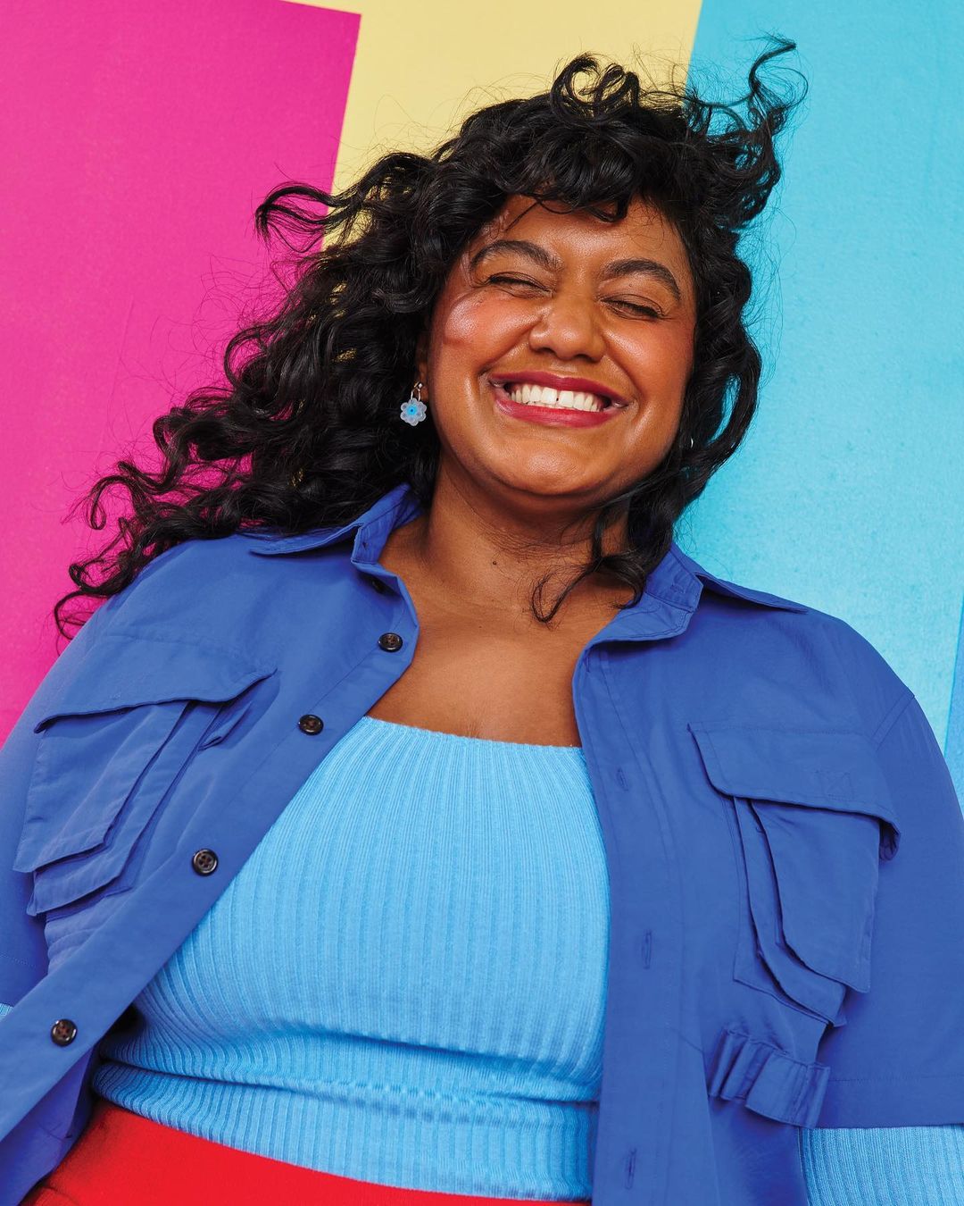 Smiling woman with curly hair poses in blue jacket against a bright backdrop of pink, yellow, and blue blocks of color indoors.