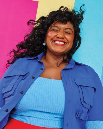Smiling woman with curly hair poses in blue jacket against a bright backdrop of pink, yellow, and blue blocks of color indoors.