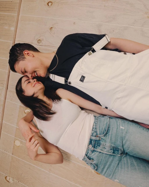 A happy couple lays close together on a light wood floor. One adult female wears a white tank top and the other wears white overalls.