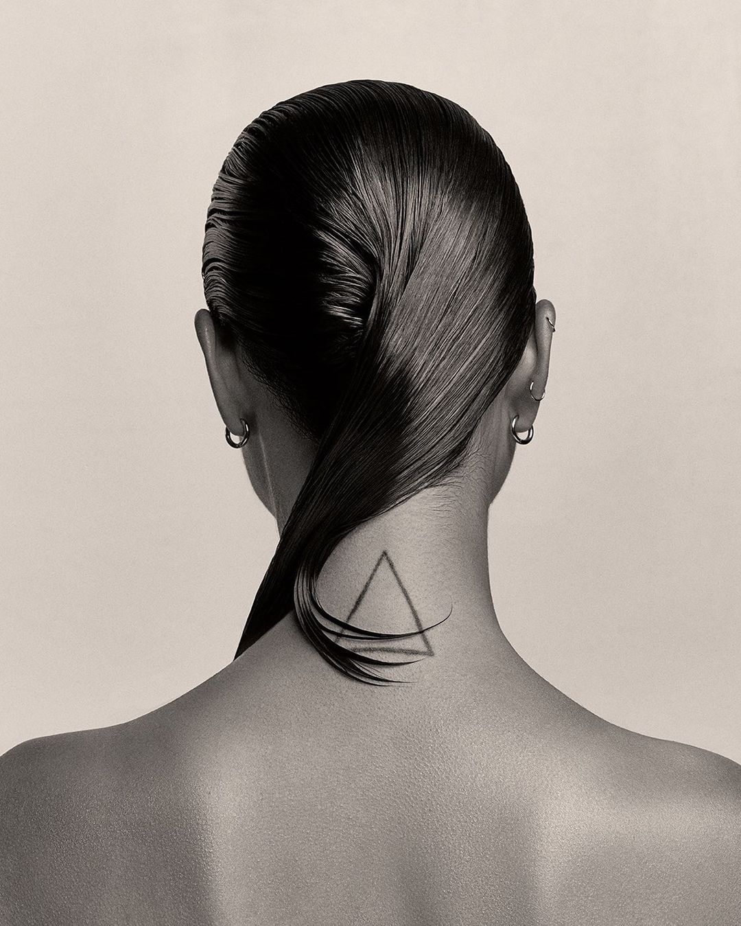 Close-up shot of an adult woman's head from behind showing slicked-back black hair, silver hoop earrings, and a triangle tattoo on her neck.