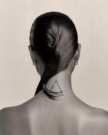 Close-up shot of an adult woman's head from behind showing slicked-back black hair, silver hoop earrings, and a triangle tattoo on her neck.