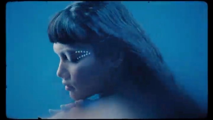 Close-up of a woman with embellished makeup, against a teal backdrop, captured with soft lighting and a shallow depth of field.