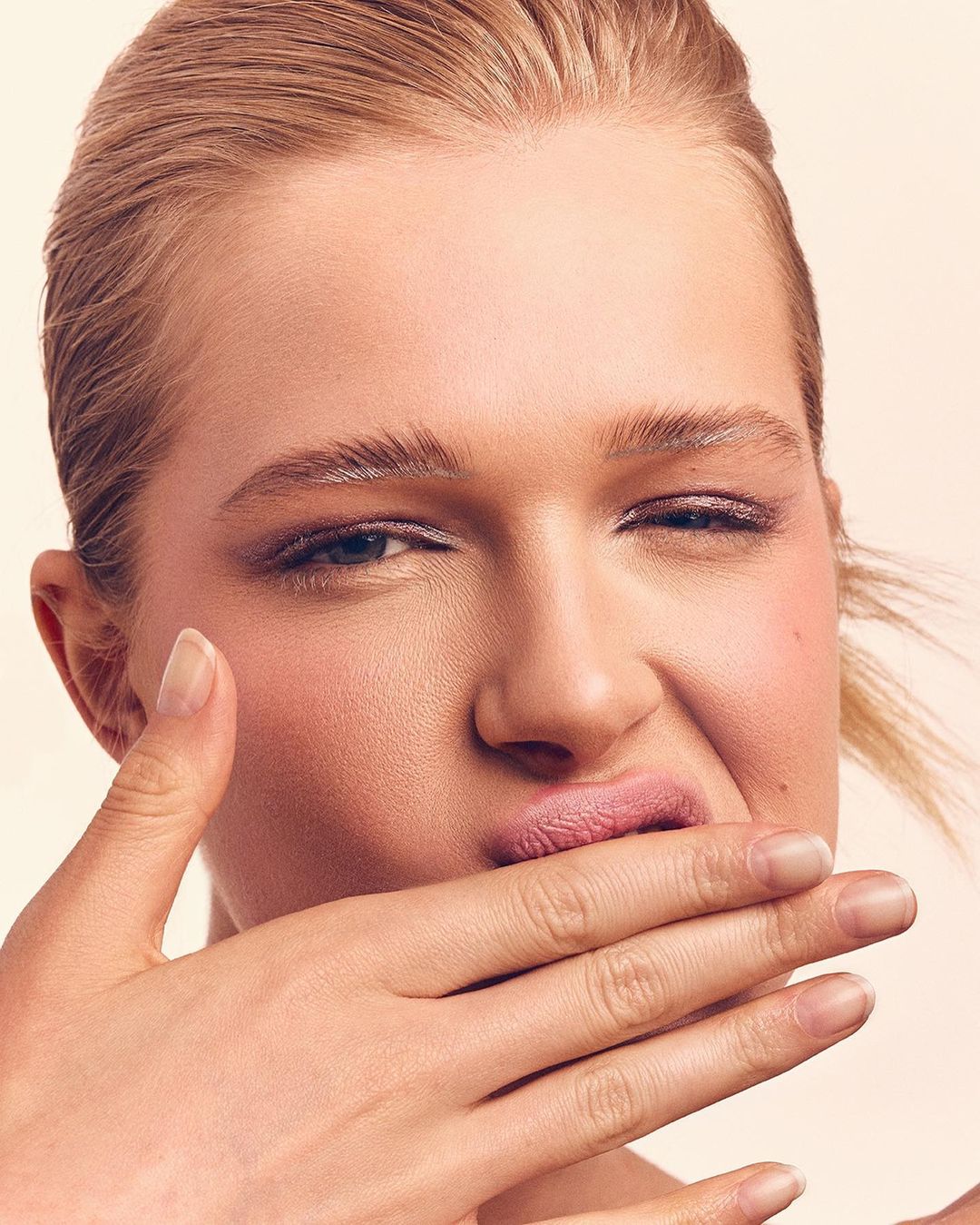 Close-up studio shot of a blond adult female model with pink blush and lipstick, covering her mouth with hand and winking against peach background.