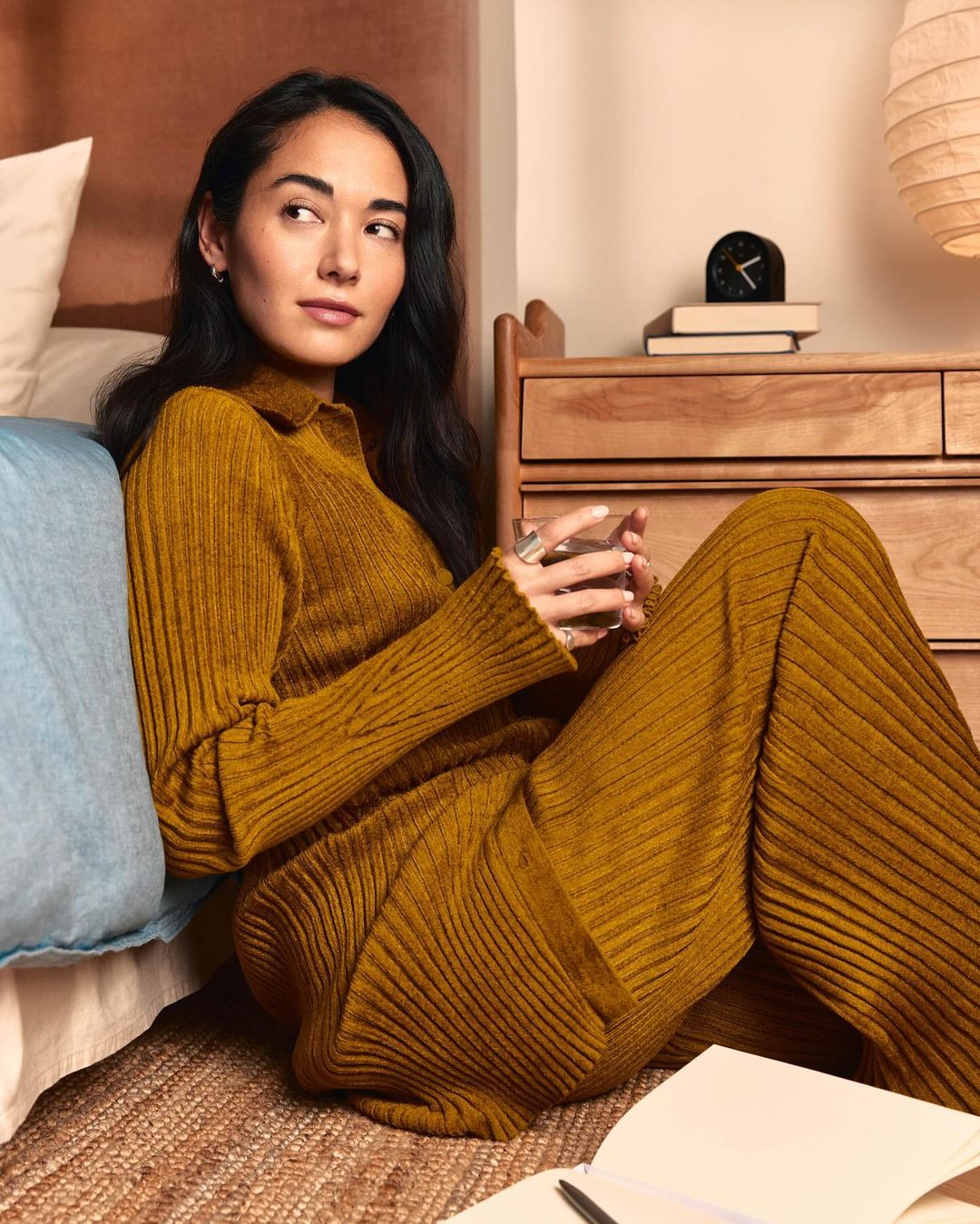 Woman in mustard-colored ribbed knitwear sits on the floor with a glass of water in a bedroom setting, casual relaxation.