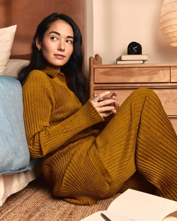 Woman in mustard-colored ribbed knitwear sits on the floor with a glass of water in a bedroom setting, casual relaxation.