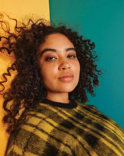 Close-up studio portrait of a woman with curly brown hair and a yellow and black plaid sweater against a yellow and teal background.