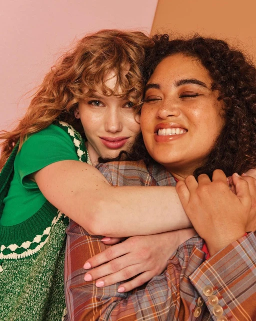 Two women embrace indoors: one with red curly hair wearing a green sweater, the other with brown curly hair in a plaid jacket.