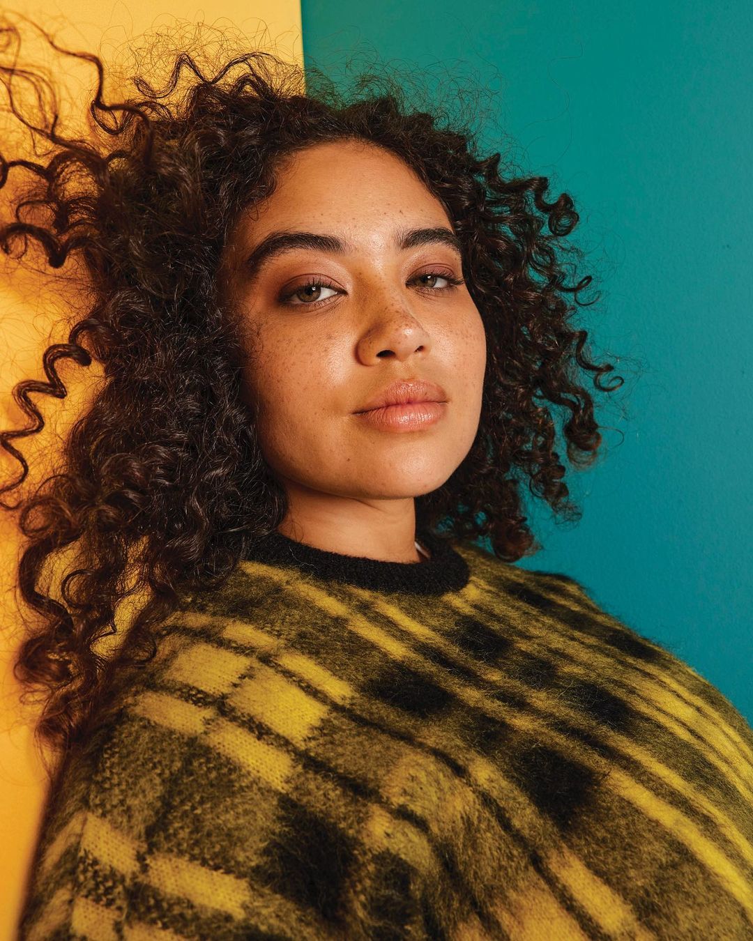 Portrait of a young woman with curly hair wearing a yellow and black plaid sweater against a blue and yellow background.
