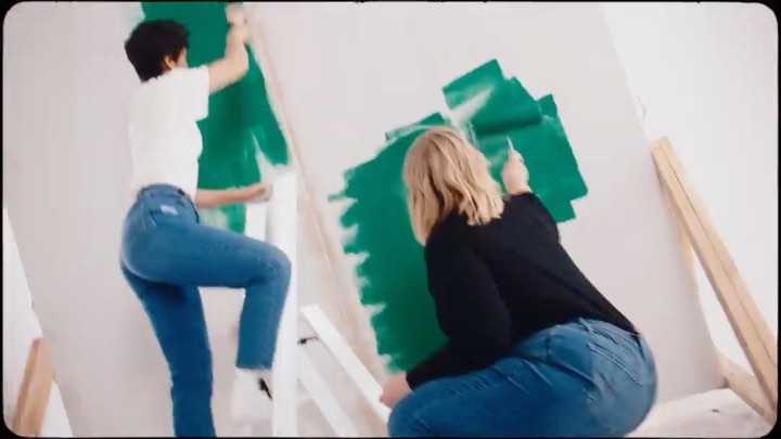 Two women paint a large white canvas with green paint indoors, using paint rollers and brushes. They are standing on ladders.