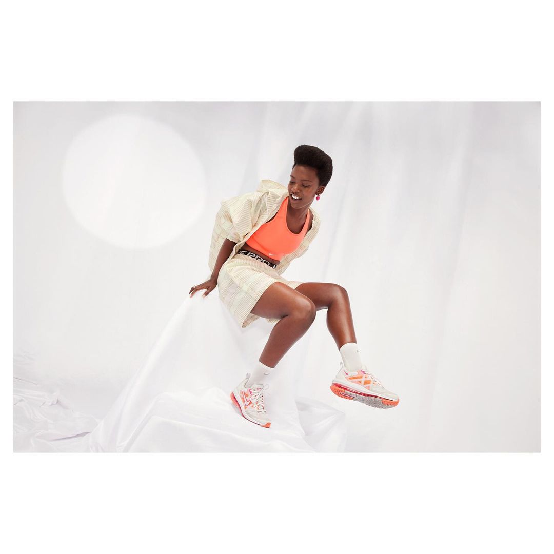 A Black woman models athletic shoes in a studio. She is sitting in a draped setting wearing athletic wear, with bright studio lighting.