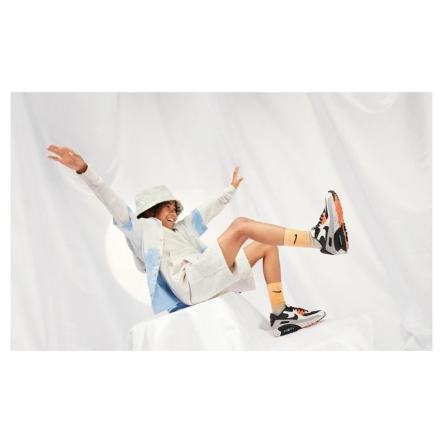 Young person balances on a white surface, wearing shorts, jacket, bucket hat, and sneakers, against a white draped backdrop.