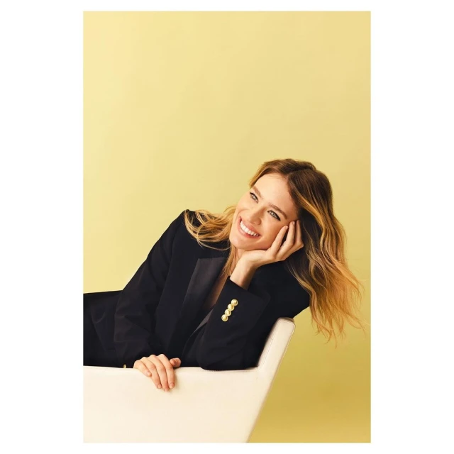 Smiling woman with blonde hair in a black blazer reclines against a white chair against a pale yellow background in a studio portrait.