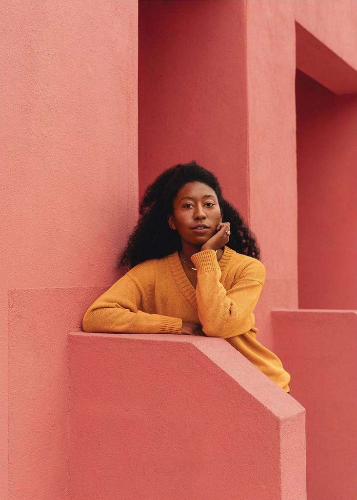 Portrait of a woman with dark curly hair in a yellow sweater leaning against a salmon-pink geometric architectural background.