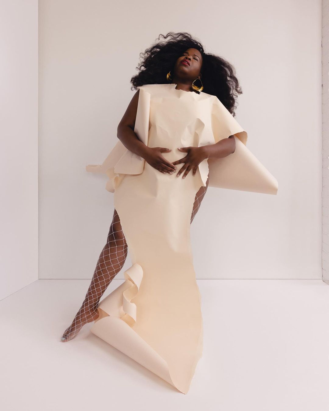 A Black woman poses in a light cream paper dress in a white studio. She is accessorized with large yellow disc earrings and wears sheer tights.