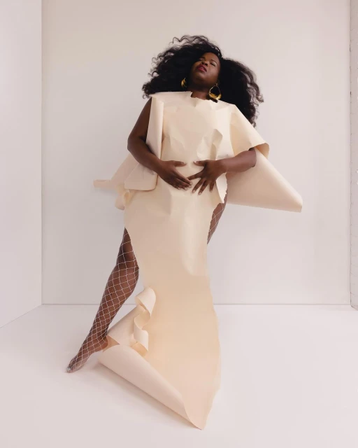 A Black woman poses in a light cream paper dress in a white studio. She is accessorized with large yellow disc earrings and wears sheer tights.
