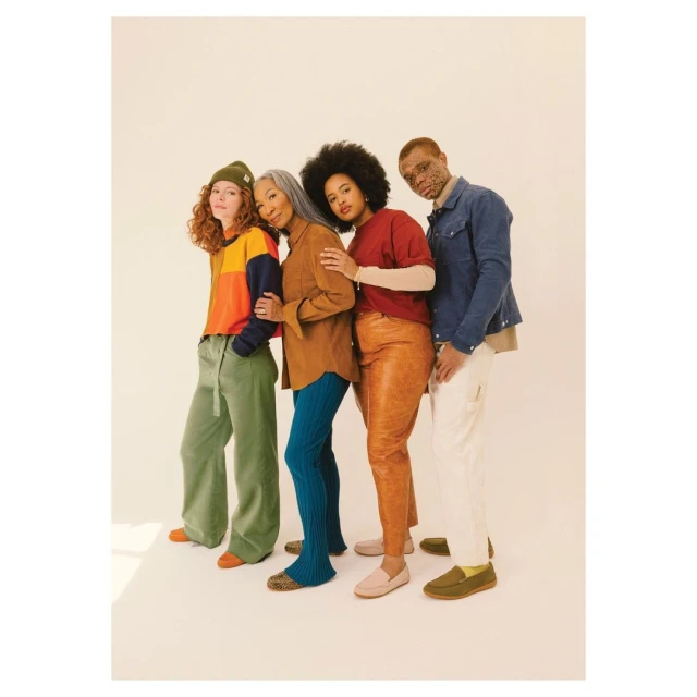 Four diverse people in casual clothing stand in a row against a cream backdrop. A warm-toned studio shot emphasizes textures and earthy colors.