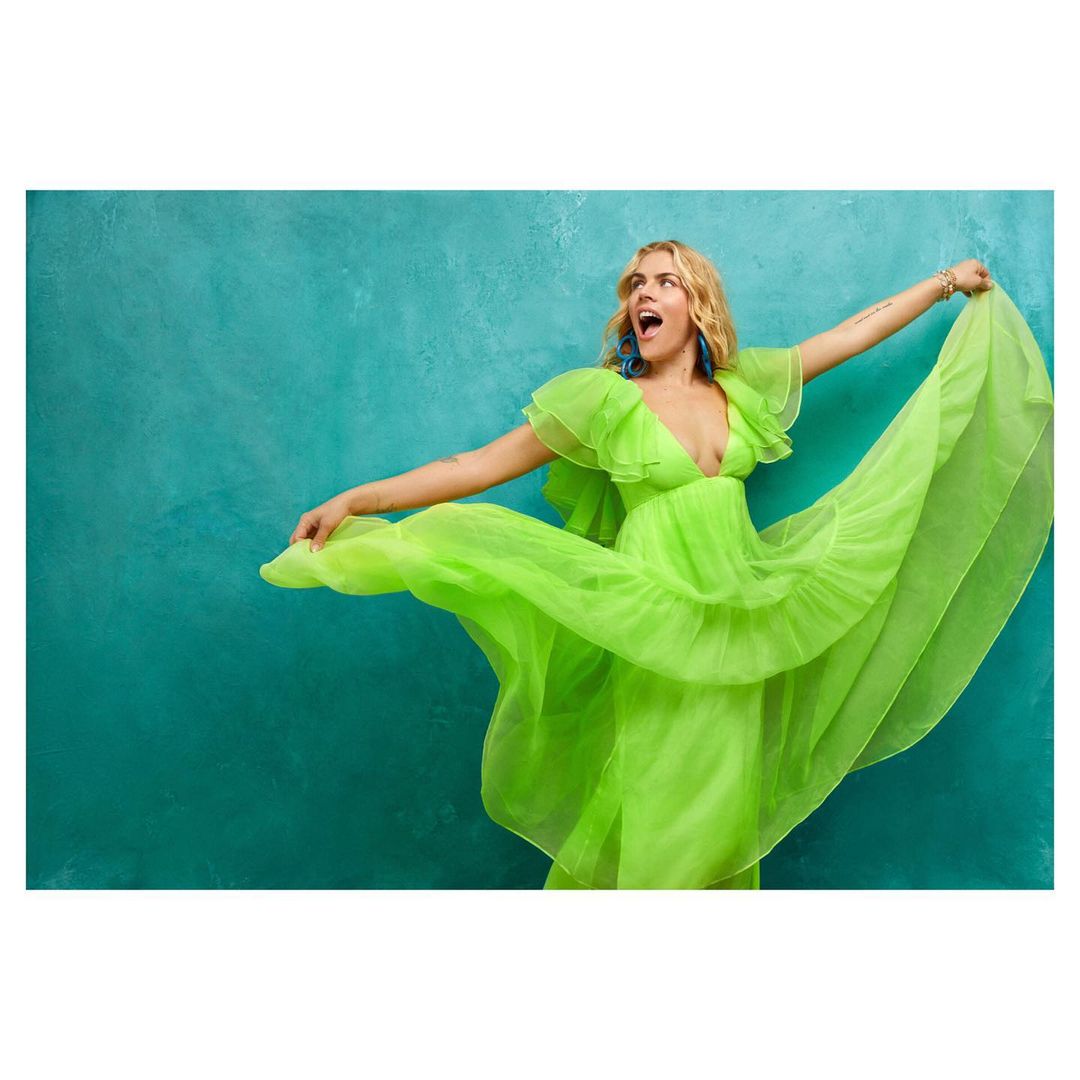 Blonde woman in a flowing neon green gown poses with arms outstretched against a textured teal background in a fashion setting.