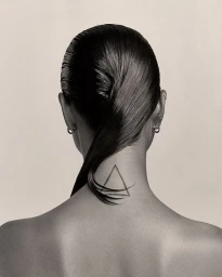 Rear view of a person with slicked-back dark hair and a geometric triangle tattoo on the back of their neck against a neutral background.