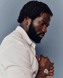 Black man with dreadlocks wearing a white shirt and leather strap watch against a grey background