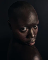 Close-up portrait of a bald Black person glancing back over their shoulder against a dark background, photographed by Grace Rivera.