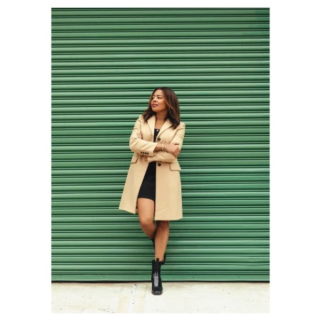 Woman in beige coat and black boots leaning against a ribbed green roller shutter door