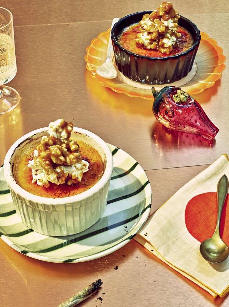 Close-up of two crème brûlées topped with walnuts, served on a copper-colored table, with a decorative pepper and a glass of water.
