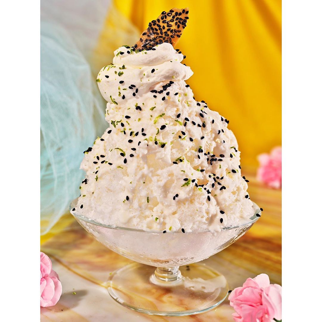 Close-up studio shot of shaved ice dessert with whipped cream, sesame seeds, and a decorative wafer in a clear glass bowl.