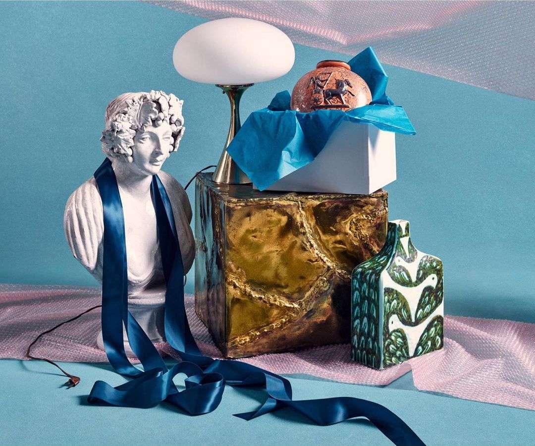 Still life featuring a white bust with a blue ribbon, a lamp, and decorative vases on a blue surface with soft pink netting background.