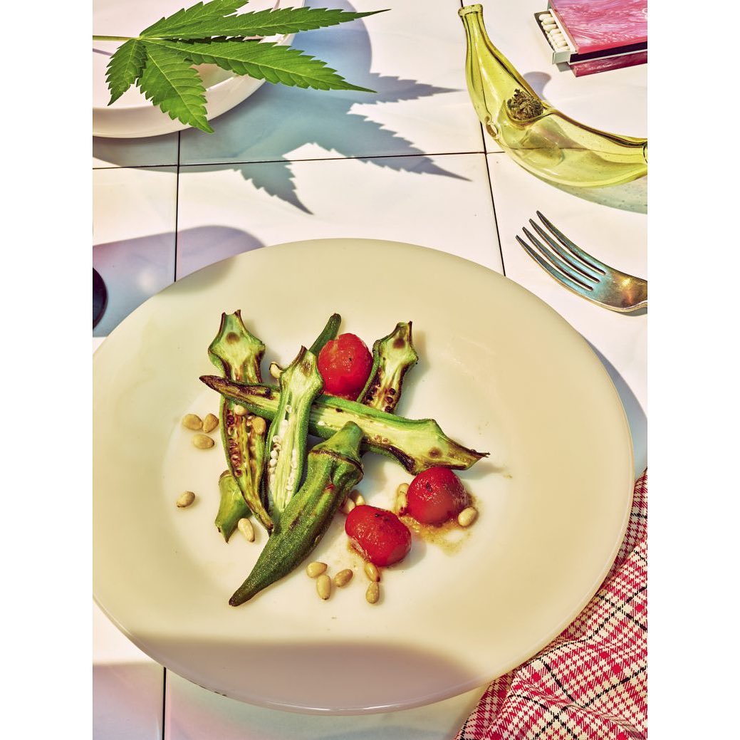Overhead shot of plate with grilled okra, red tomatoes, and pine nuts; cannabis leaf and glass banana in background on tiled surface.