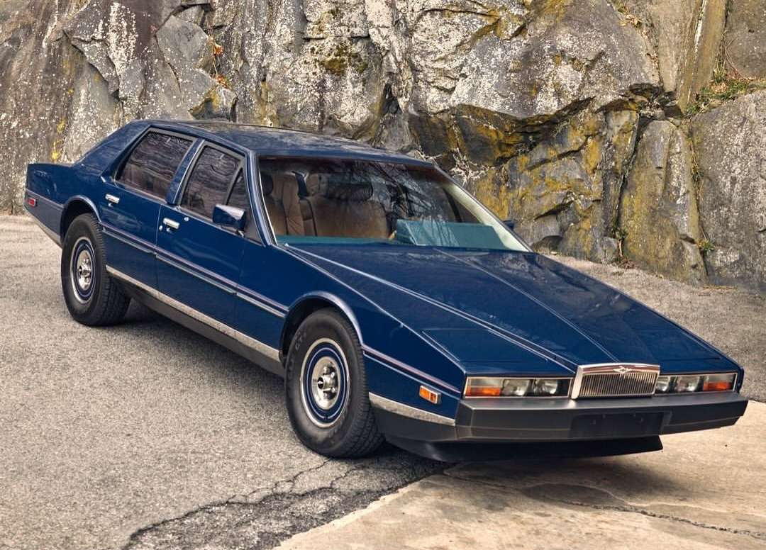 Dark blue Aston Martin Lagonda sedan parked on asphalt against a background of large rocks and sparse vegetation on a cloudy day.