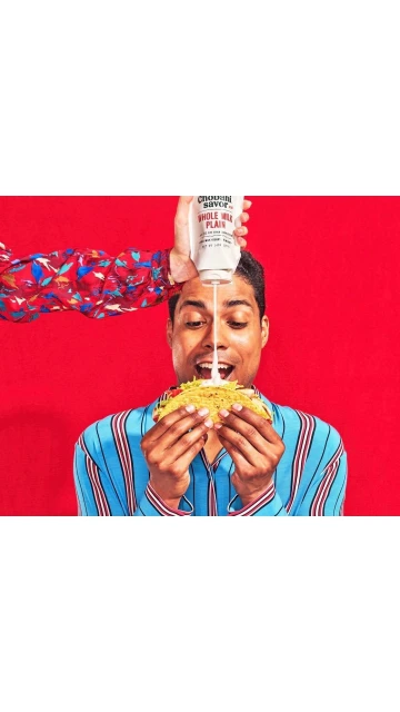 Man holding a taco with sauce being poured on it from above. He is wearing a blue striped shirt with a red background.