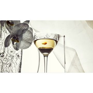 A glass of clear liquid sits beside a black and white orchid, highlighted by a touch of orange, on a soft white background.