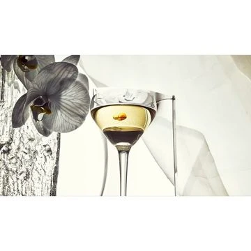 A glass of clear liquid sits beside a black and white orchid, highlighted by a touch of orange, on a soft white background.