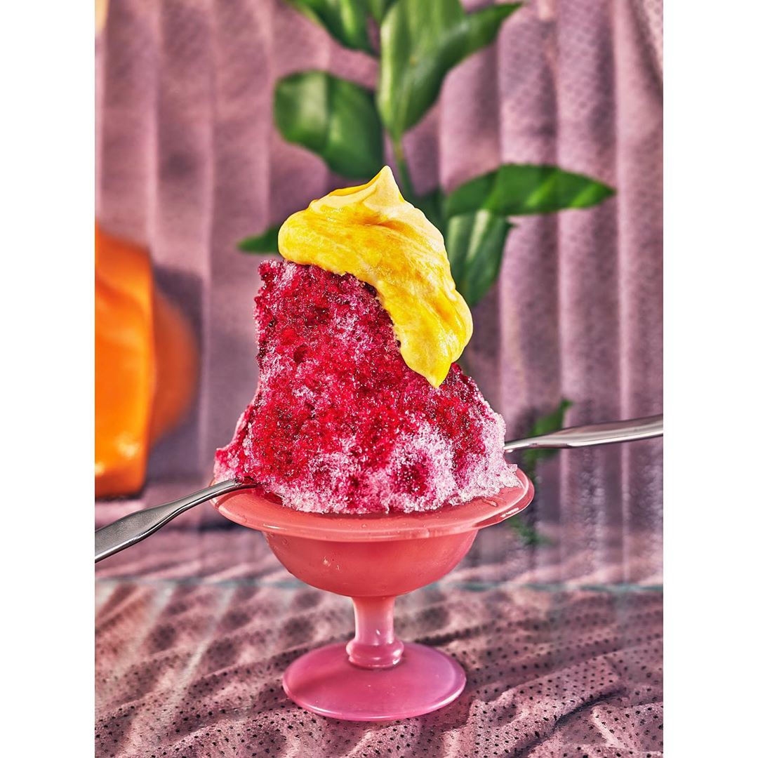 Red shaved ice in a pink stemmed bowl topped with yellow cream and two spoons, against a decorative background with greenery.