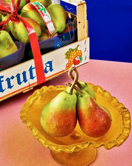 Close-up still life of a golden bowl holding three pears, with a wooden crate of pears in the background against pink and blue.
