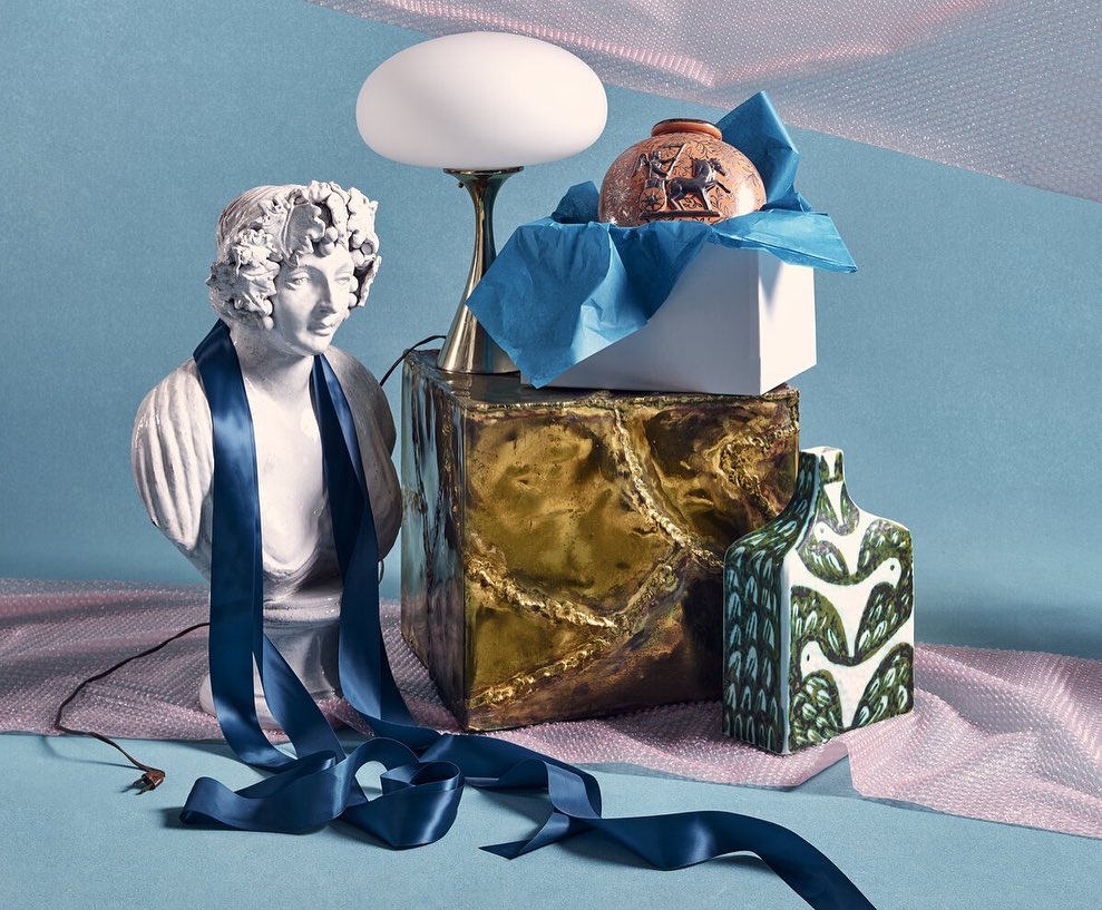 Still life featuring a bust with a blue ribbon, a lamp on a metallic box, a small vase, and a patterned ceramic vase on a pink fabric.