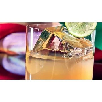 Close-up shot of an amber-colored cocktail in a clear glass with a large ice cube and a lime wedge garnish, resting on a reflective surface.